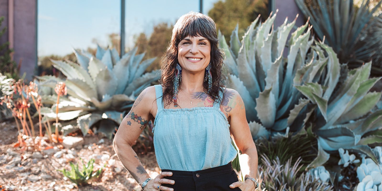 Erika Wright standing confidently outdoors among large agave plants, smiling with hands on hips, wearing a light blue top and long beaded earrings.