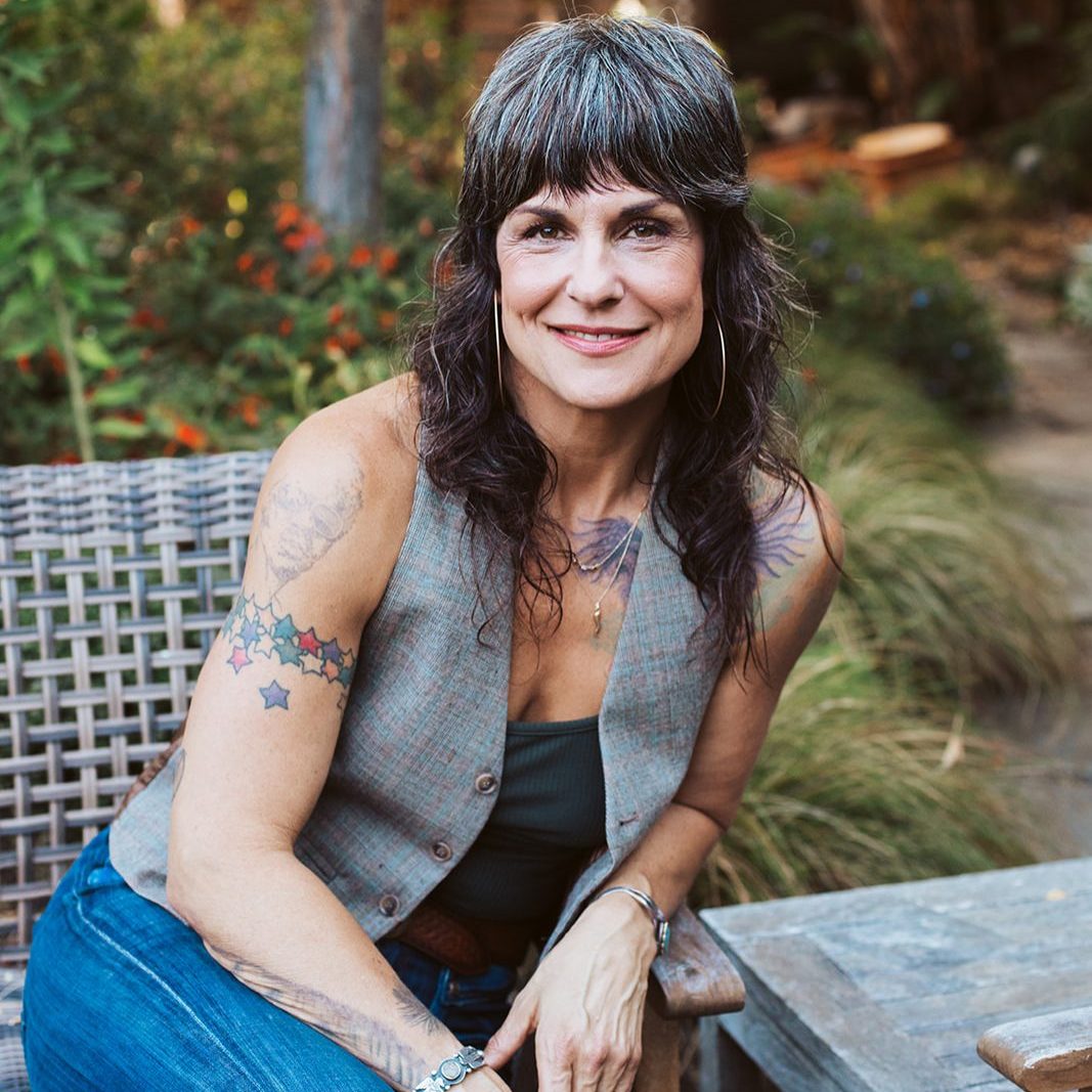 Smiling woman with dark gray hair and tattoos sits on a wicker chair in a garden patio outdoors.